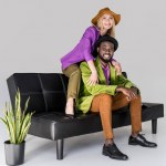 Cheerful multicultural fashionable couple in hats sitting on black sofa on grey background