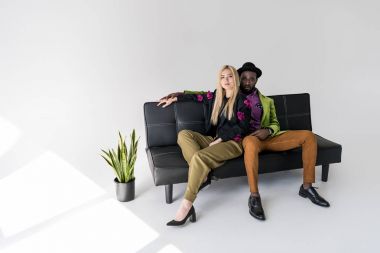 multicultural fashionable couple resting on black sofa on grey backdrop clipart