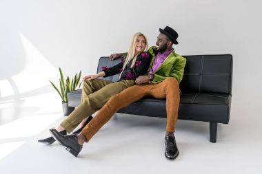 cheerful multicultural fashionable couple resting on black sofa on grey background clipart