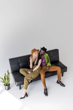 high angle view of smiling multicultural fashionable couple sitting on black sofa  clipart