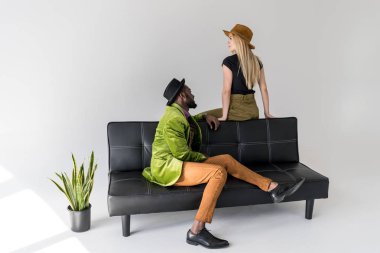 multicultural fashionable couple in hats on black sofa on grey backdrop clipart