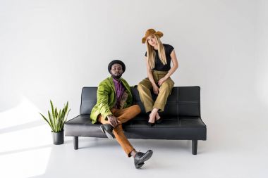 young multicultural fashionable couple sitting on black sofa on grey backdrop clipart