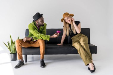 african american fashionable man presenting flower to smiling caucasian girlfriend on black sofa clipart