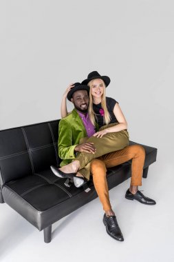 kanepede oturan ve gri kameraya gülümseyen şapkalar moda çok ırklı çiftin yüksek açılı görünüş