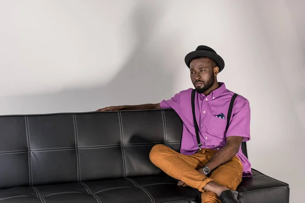 帽子灰色の背景に黒いソファの上で物思いにふけるアフリカ系アメリカ人のスタイリッシュな男 — ストック写真