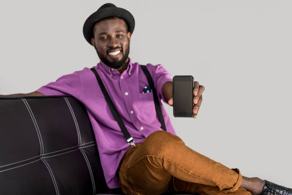 selective focus of smiling african american stylish man showing smartphone with blank screen on black sofa isolated on grey