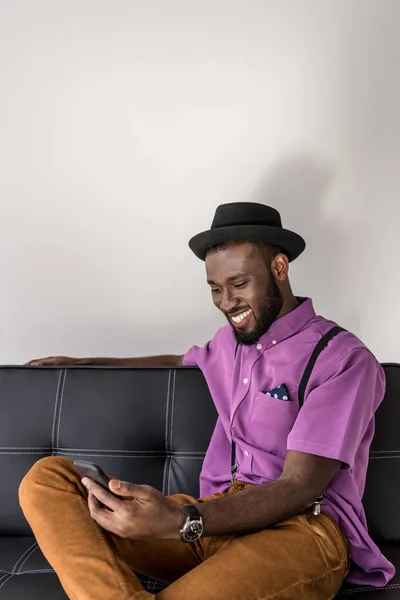 灰色の背景に黒いソファにスマート フォンを使用してアフリカ系アメリカ人のスタイリッシュな男の笑みを浮かべてください — ストック写真