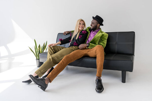 cheerful multicultural fashionable couple resting on black sofa on grey background