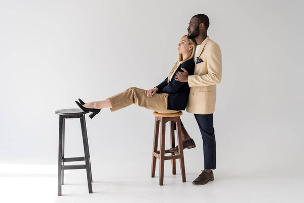 side view of young african american man embracing beautiful stylish girlfriend sitting on chair and looking away on grey