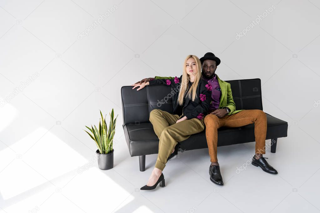 multicultural fashionable couple resting on black sofa on grey backdrop