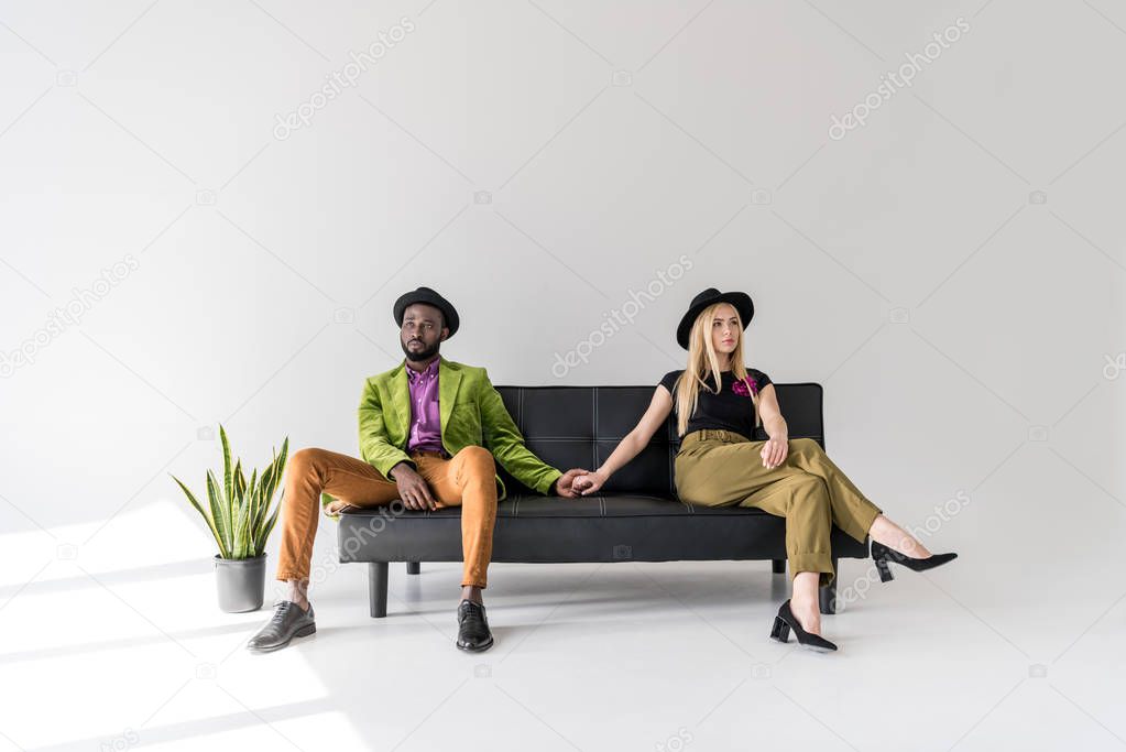 stylish young multiethnic couple holding hands while sitting on sofa and looking away on grey 