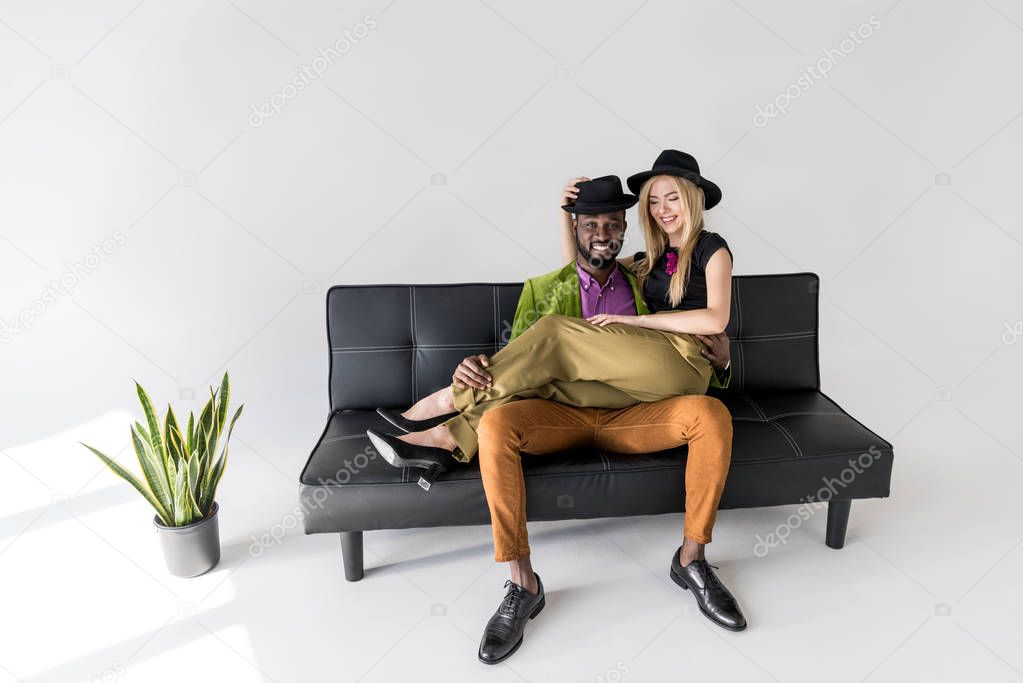 happy young multiethnic couple in stylish clothes sitting on sofa and smiling at camera on grey  