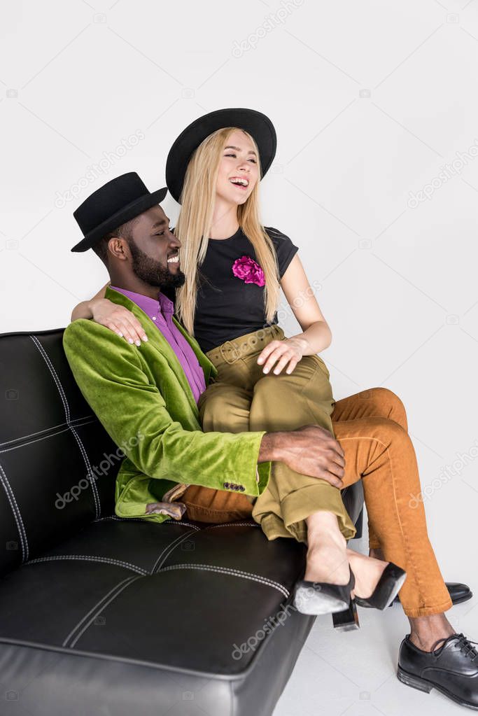 happy young multiethnic couple in stylish clothes sitting on sofa and looking away on grey  