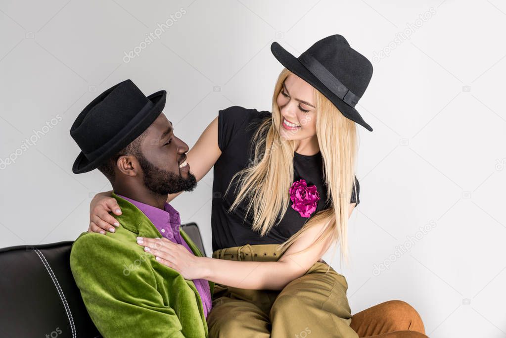 beautiful stylish young multiethnic couple in black hats smiling each other isolated on grey