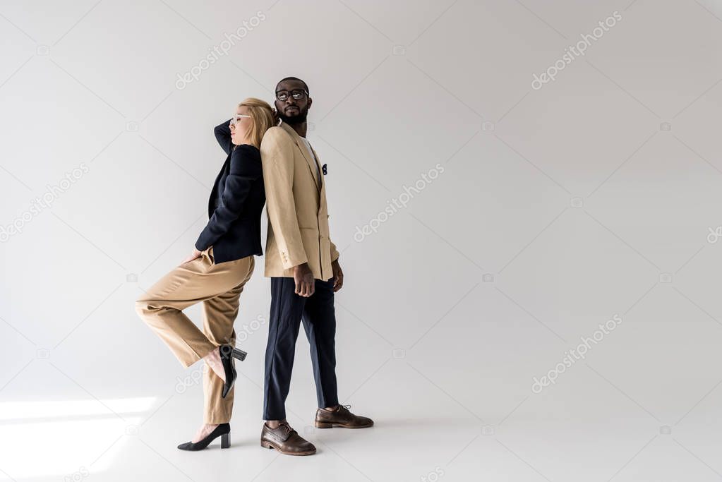 side view of fashionable young multiethnic couple posing back to back on grey