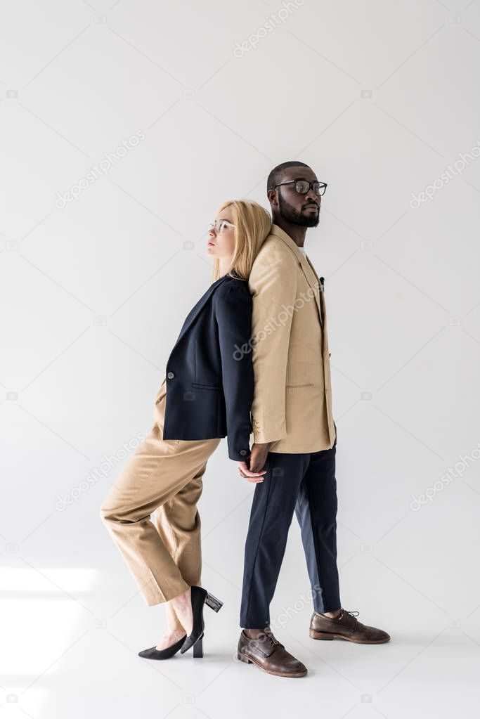 side view of stylish young multiethnic couple standing back to back and holding hands on grey