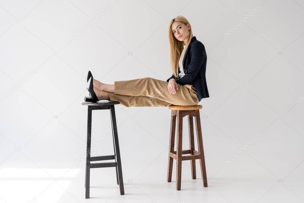 side view of beautiful young woman sitting on stools and looking at camera on grey 