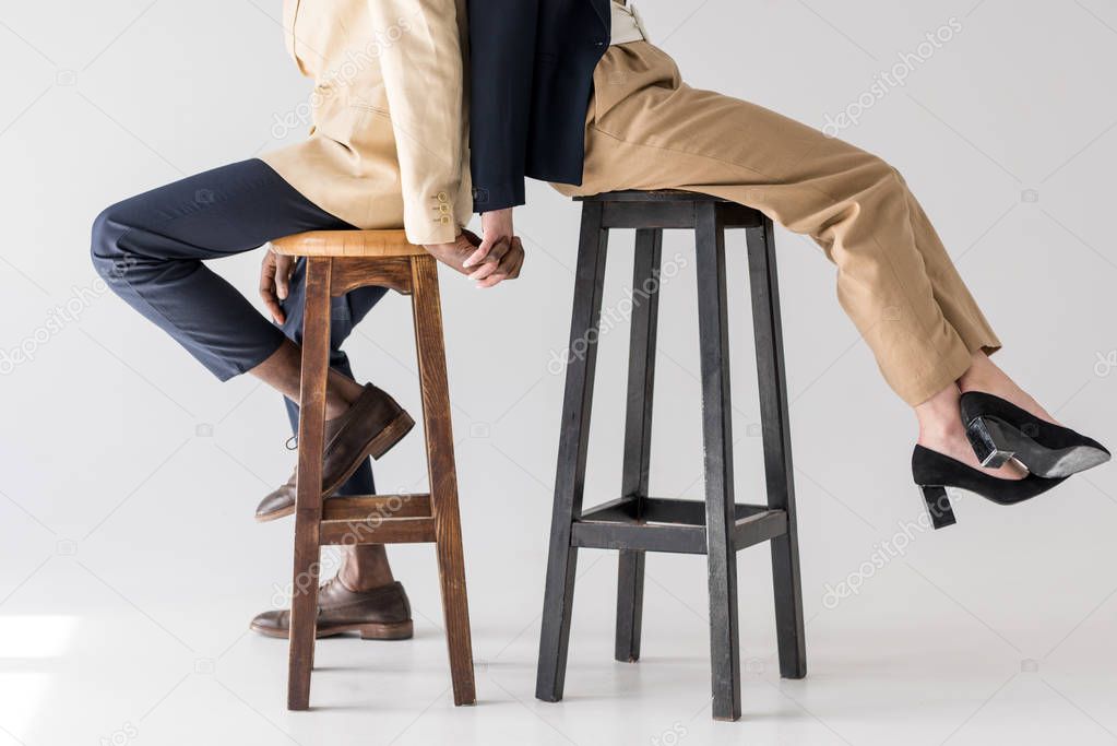 cropped shot of multiethnic couple sitting back to back on stools and holding hands on grey