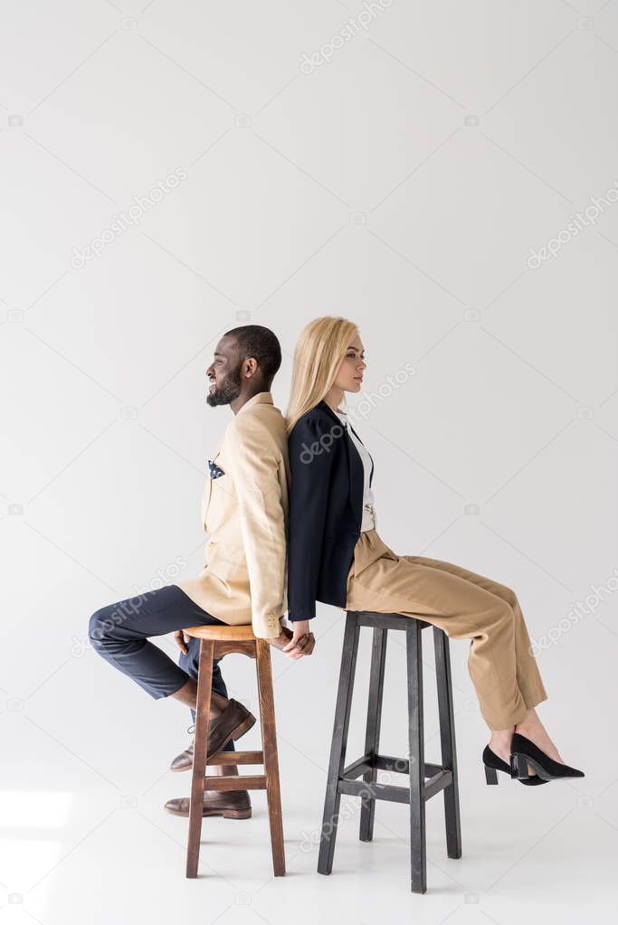 side view of fashionable young multiethnic couple sitting back to back on stools and holding hands on grey