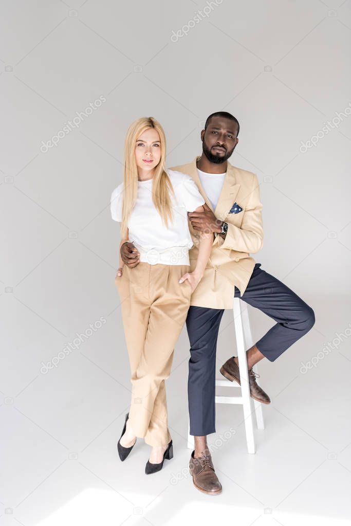 full length view of stylish young multiethnic couple looking at camera on grey 