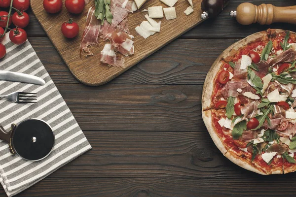 Flat lay with arranged italian pizza, cutlery and various ingredients on wooden surface — Stock Photo