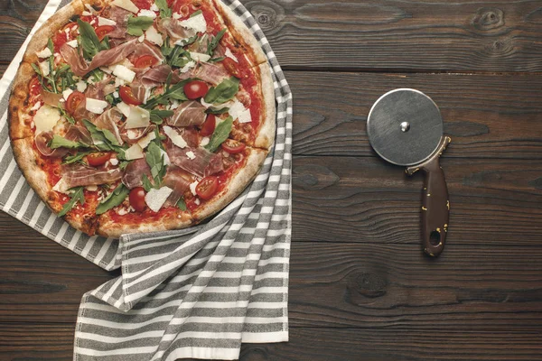 Top view of italian pizza on tablecloth and pizza cutter on wooden surface — Stock Photo