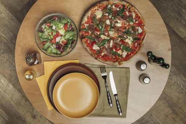 Vista superior de la disposición de platos vacíos, pizza y ensalada en la mesa de madera - foto de stock