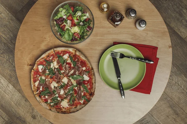 Vista superior de la disposición de ensalada en tazón, pizza italiana y vajilla en mesa de madera - foto de stock