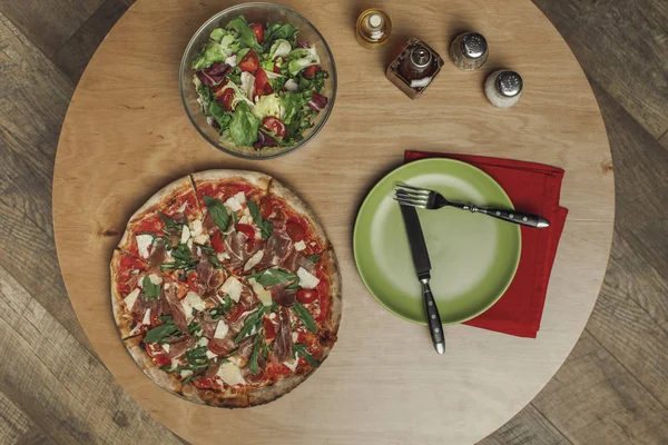 Top view of arrangement of salad in bowl, italian pizza and dinnerware on wooden table — Stock Photo