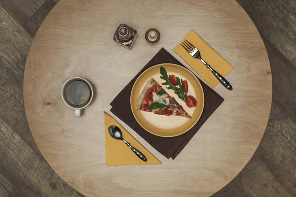 Flat lay com peça arranjada de pizza na placa, xícara de chá e talheres em mesa de madeira — Fotografia de Stock