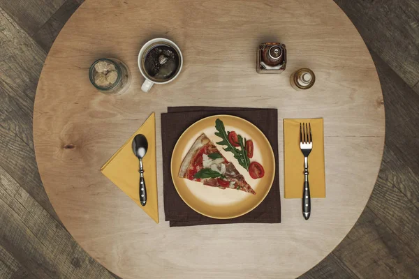 Flat lay com peça arranjada de pizza na placa, xícara de chá e talheres em mesa de madeira — Fotografia de Stock