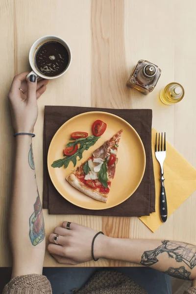 Частичный вид женщины, сидящей за столом с кусочком пиццы на тарелке и чашкой кофе — стоковое фото