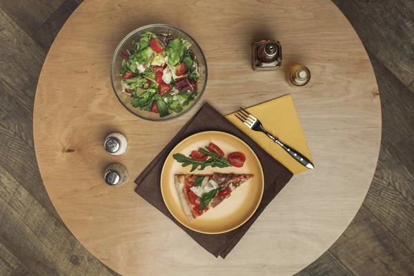 Vista superior de pedaço de pizza na placa, salada na tigela e especiarias na superfície de madeira — Fotografia de Stock