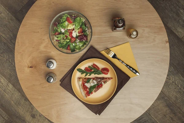 Vue du dessus du morceau de pizza sur assiette, salade dans un bol et épices sur la surface en bois — Photo de stock