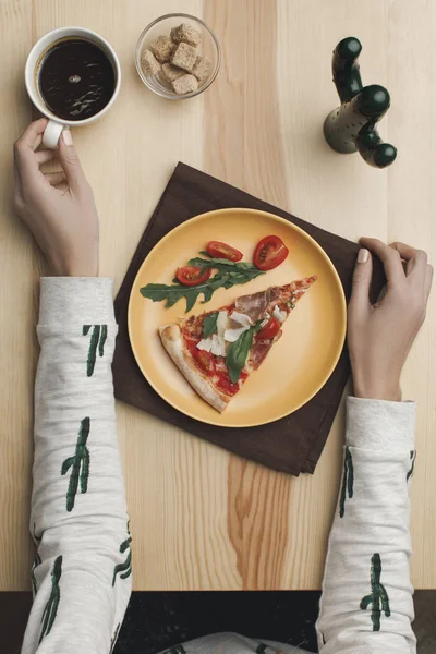 Partial view of woman sitting at table with pizza piece on plate and cup of coffee — Stock Photo