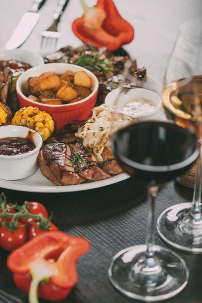 Plate with sauces, roasted potatoes, grilled meat with vegetables and glasses of wine on table — Stock Photo