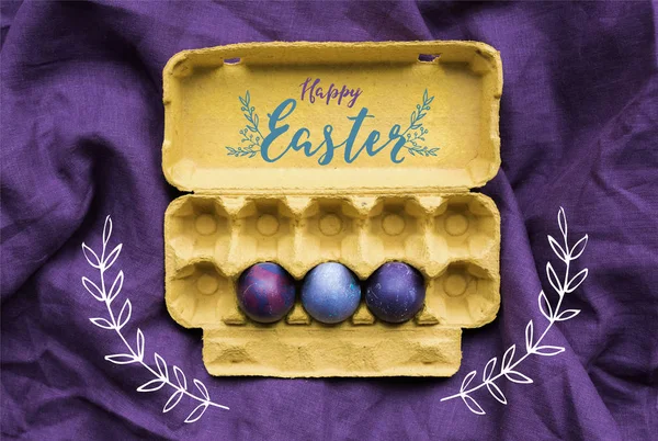 Three Easter eggs in carton  with happy easter lettering on textile background — Stock Photo