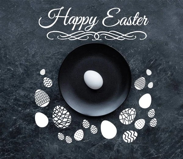 White egg on black plate  with happy easter lettering and drawn eggs on dark background — Stock Photo