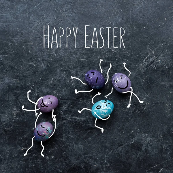 Colorful Easter eggs with drawn faces and happy easter lettering on dark background — Stock Photo