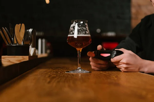Vaso de cerveza fría fresca y persona jugando con joystick detrás en el pub - foto de stock