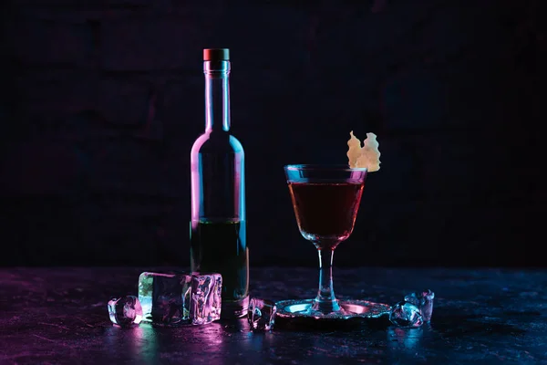 Glass of alcohol cocktail, bottle of liquor and ice cubes on dark surface — Stock Photo