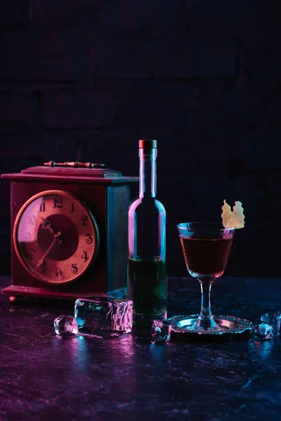 Vaso de cóctel de alcohol, botella de licor y reloj vintage en la superficie oscura - foto de stock