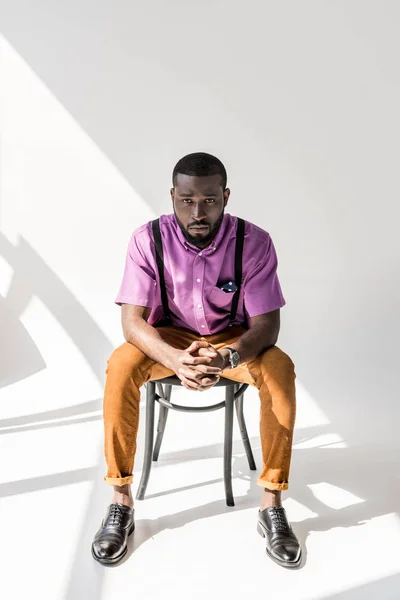 Молодий афроамериканський чоловік у стильному одязі з руками в замку, сидячи на стільці на сірому фоні — стокове фото