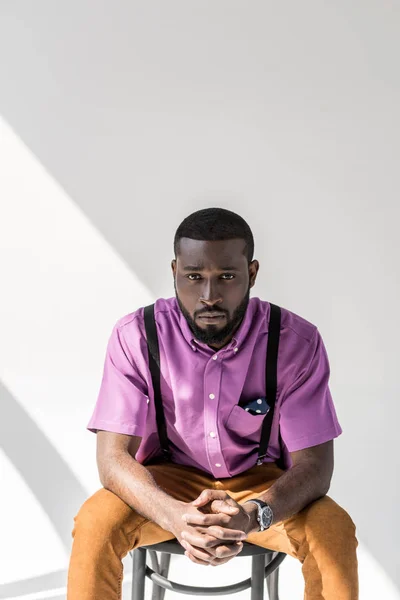 Retrato de homem afro-americano em roupas da moda com as mãos na fechadura sentado na cadeira em pano de fundo cinza — Fotografia de Stock