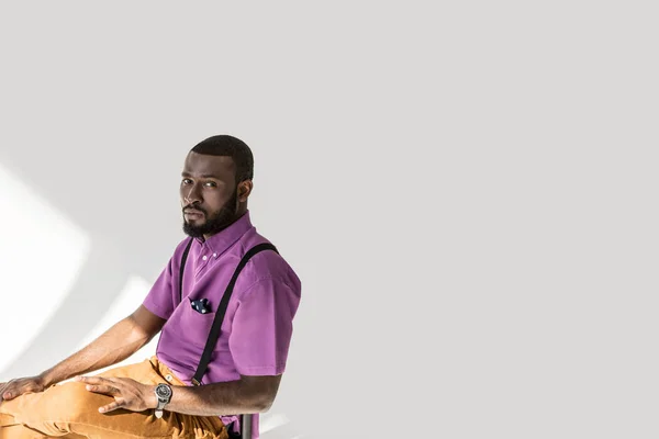 Seitenansicht eines afrikanisch-amerikanischen Mannes in modischer Kleidung, der auf einem Stuhl vor grauem Hintergrund sitzt — Stockfoto