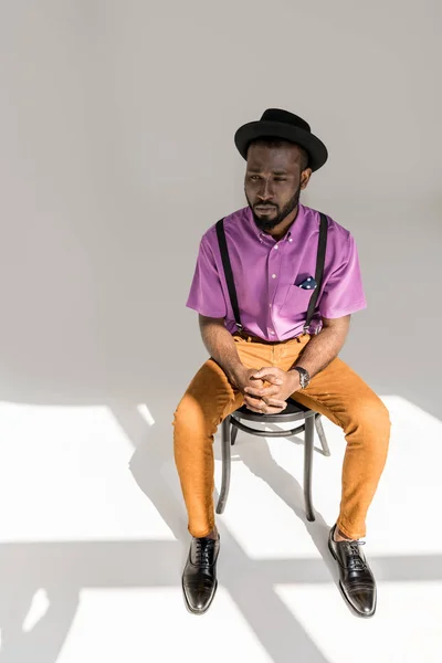 Homem americano africano pensativo em roupas elegantes e chapéu sentado na cadeira em pano de fundo cinza — Fotografia de Stock