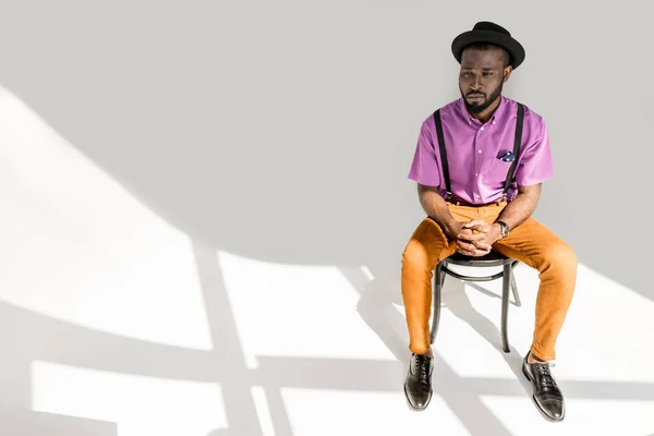 Пенсійний афроамериканський чоловік у стильному одязі та капелюсі сидить на стільці на сірому фоні — стокове фото