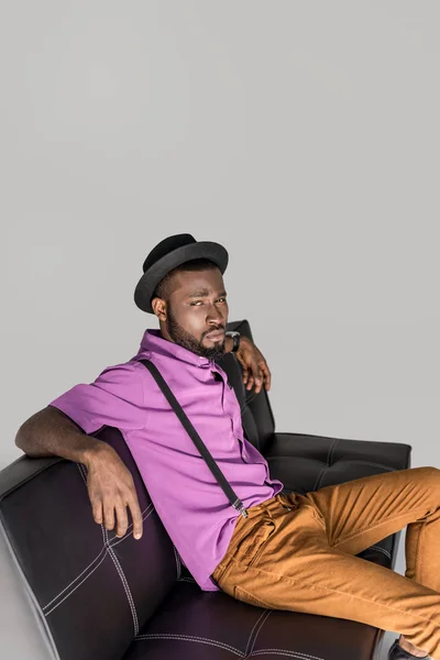 Вид сбоку африканского мужчины в стильной шляпе, сидящего на черном диване, изолированном серым — стоковое фото