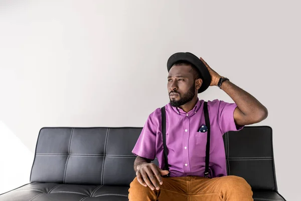 Портрет задумчивого африканского стильного американца в шляпе на черном диване на сером фоне — стоковое фото