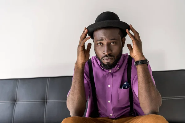 Retrato de afro americano elegante homem de chapéu no sofá preto isolado no cinza — Fotografia de Stock